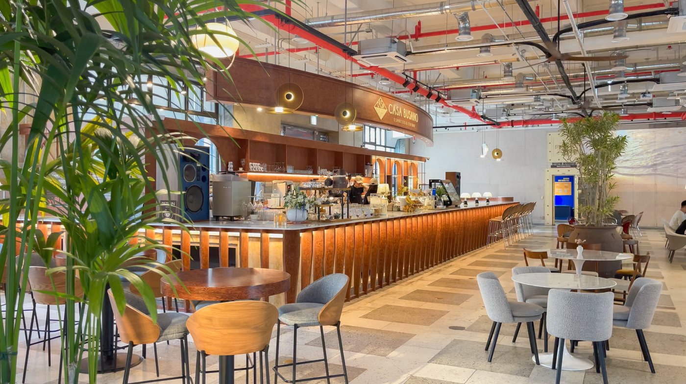 Cafe counter, tables and indoor plants beneath a CASA BUSANO sign.