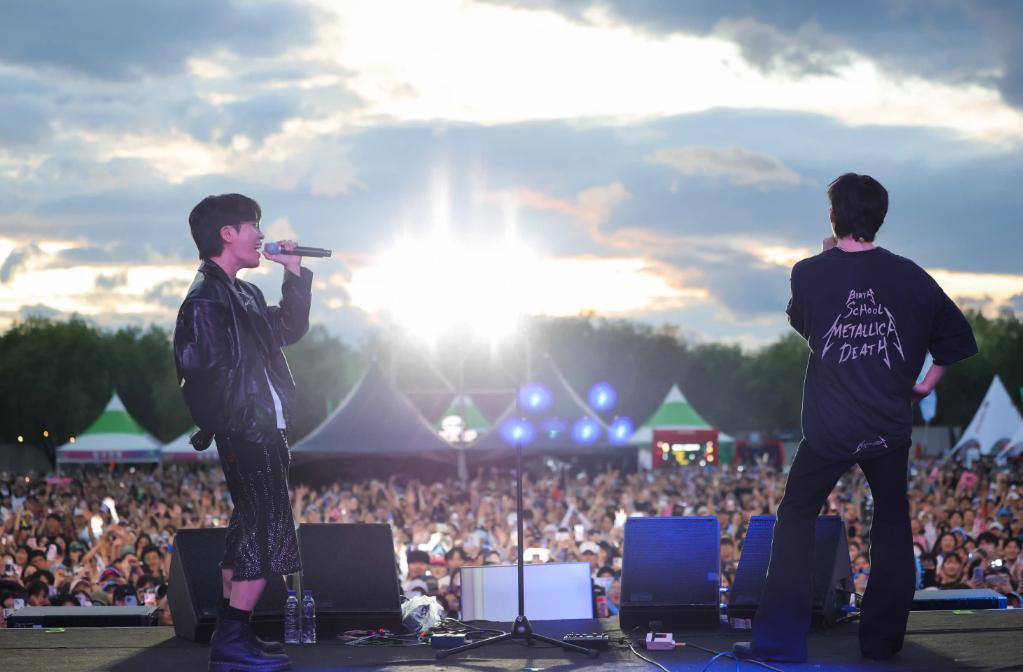 Two performers on an outdoor stage facing a festival crowd near sunset