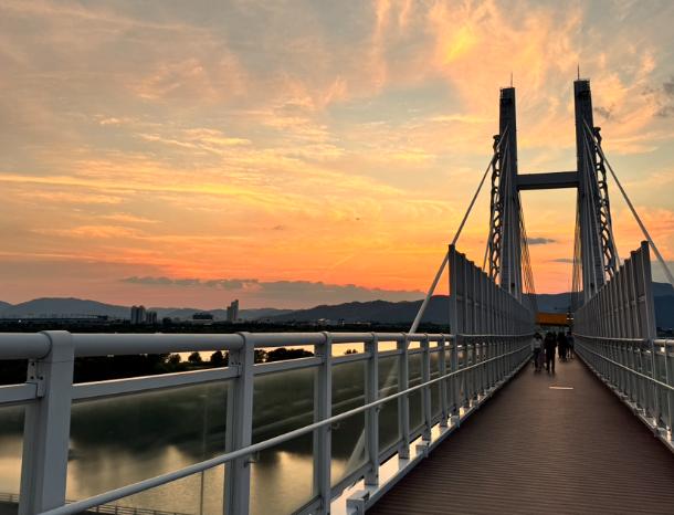 Pedestrian bridge over the riverside landscape beneath an orange sunset
