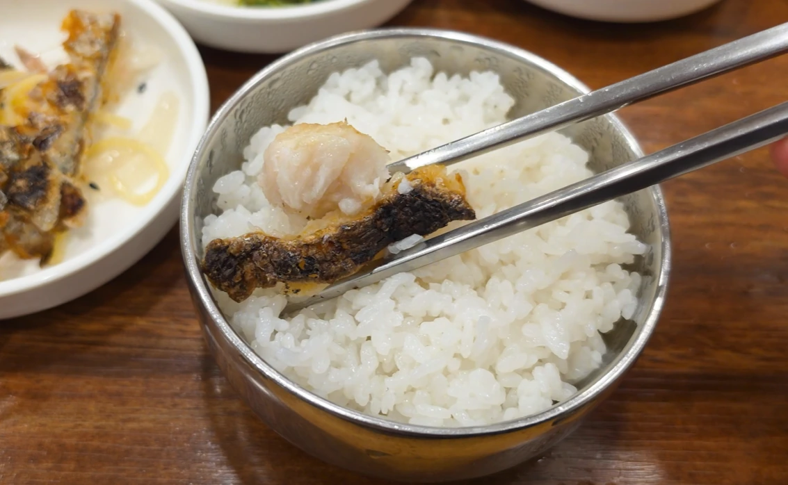 A piece of grilled cutlassfish held in metal chopsticks above a bowl of white rice.