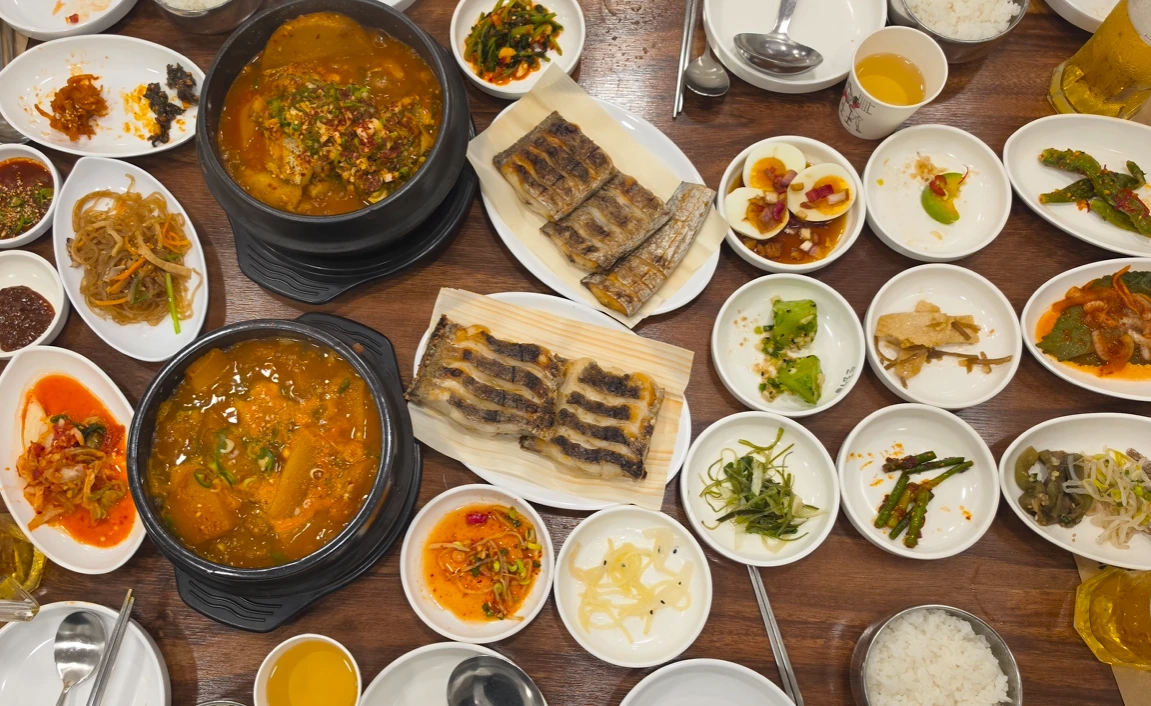 Grilled cutlassfish on plates beside two bowls of stew and assorted Korean side dishes.