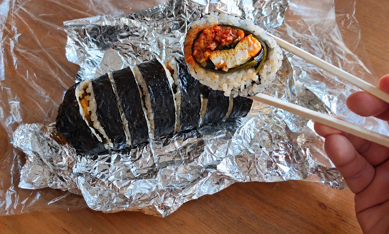 Sliced gimbap on aluminum foil, with one piece held in chopsticks to show the filling.