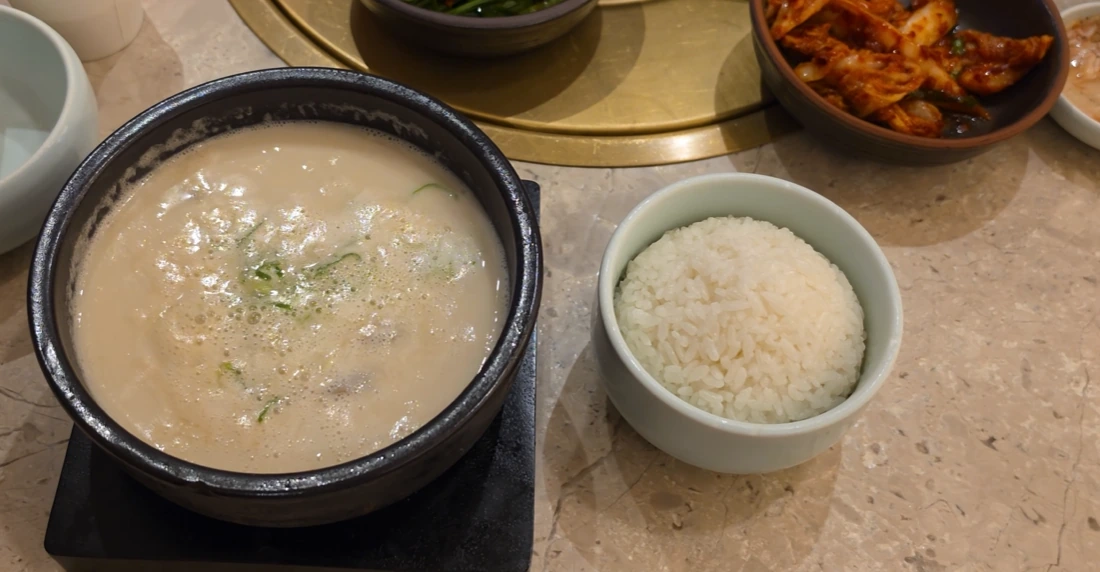 A black bowl of pale pork broth beside white rice and kimchi at Geodae Dwaeji Gukbap.