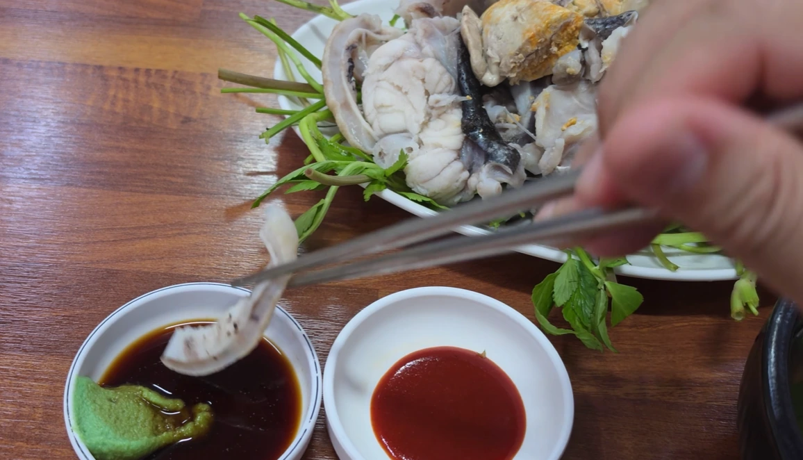 Chopsticks holding a piece of monkfish above soy sauce and wasabi, with the steamed platter behind.