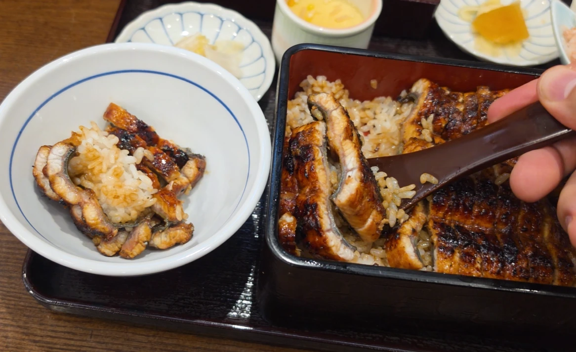 A wooden spoon transferring grilled eel and rice from a lacquered box into a smaller bowl.