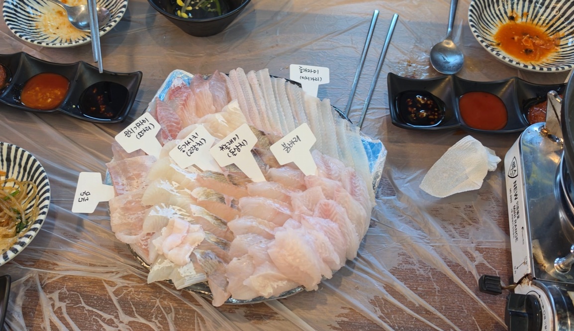 A platter of sliced raw fish with Korean species labels, surrounded by dipping sauces and side plates.