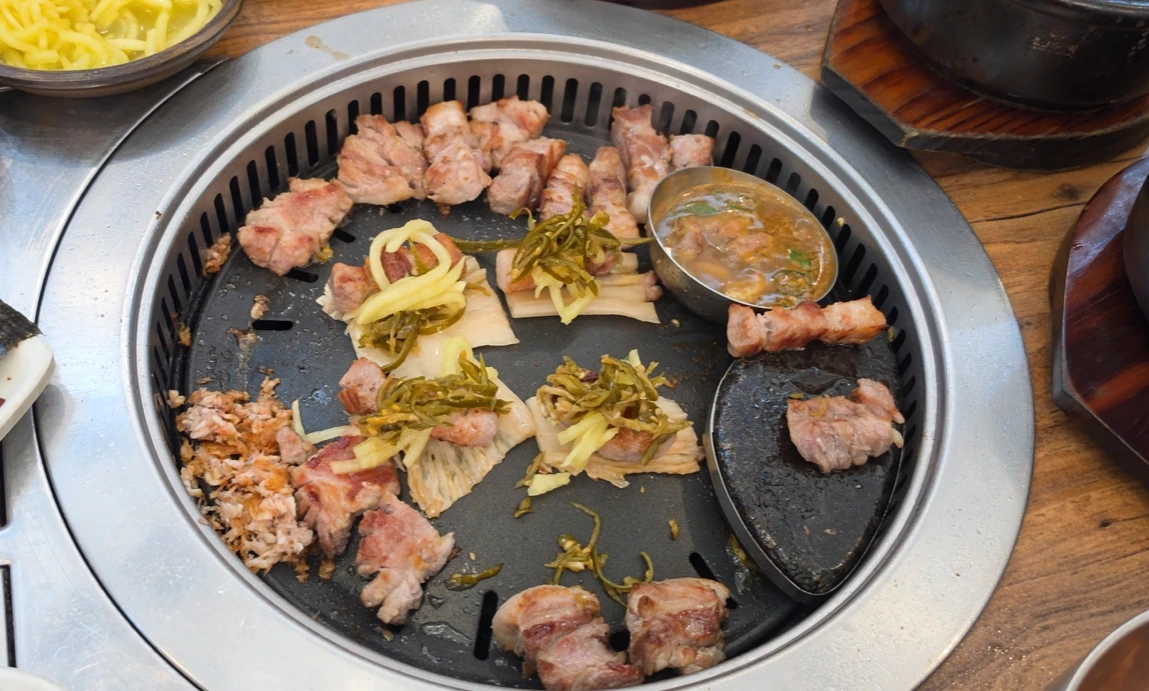 Browned pork pieces on a tabletop grill, with several bites topped with pale and green pickled vegetables.