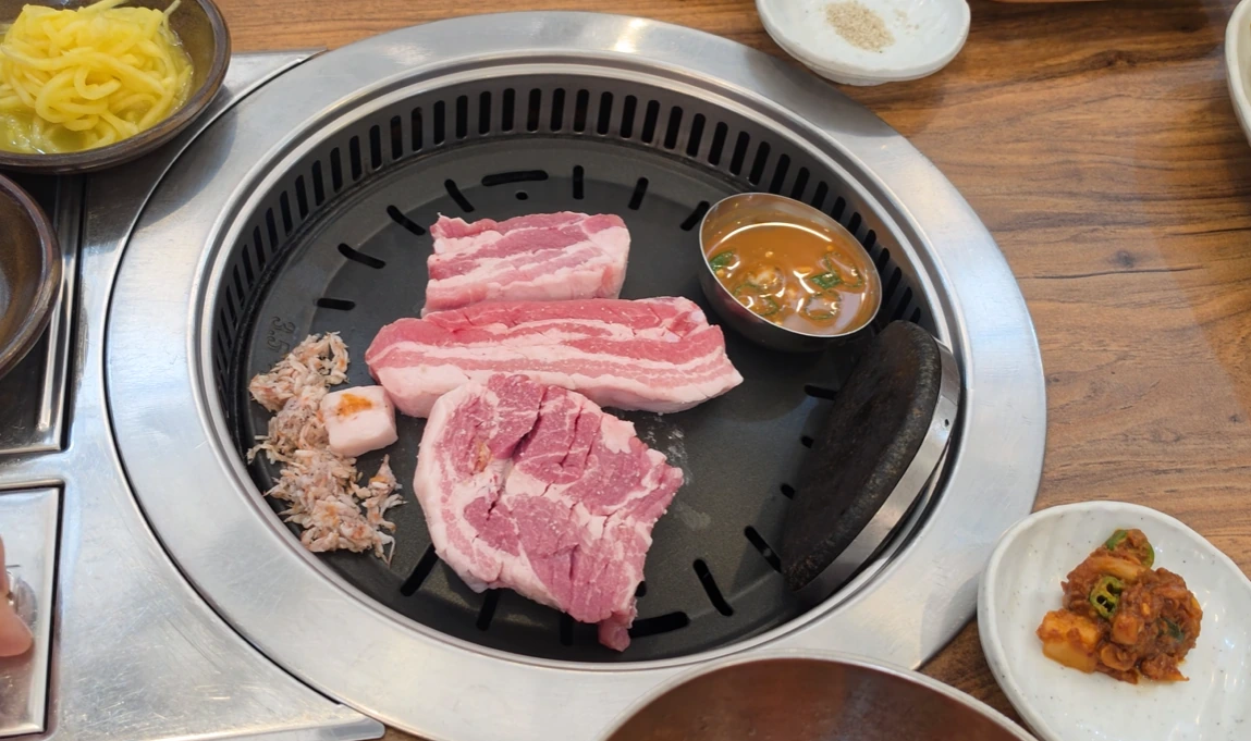 Thick slices of raw pork belly and pork neck on a round tabletop grill with a small sauce bowl.