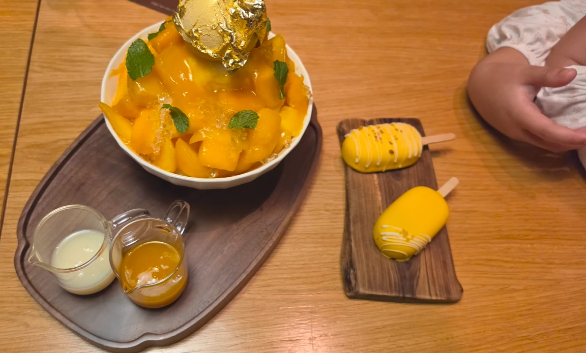 A large bowl of mango-topped shaved ice with mint and a gold-colored garnish, small sauce jugs and two yellow frozen treats.