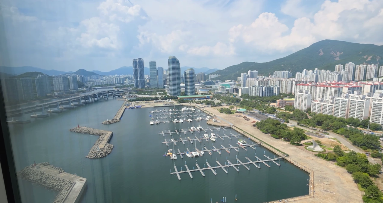 A marina with moored yachts, waterfront towers and green hills seen through a window at Park Hyatt Busan.