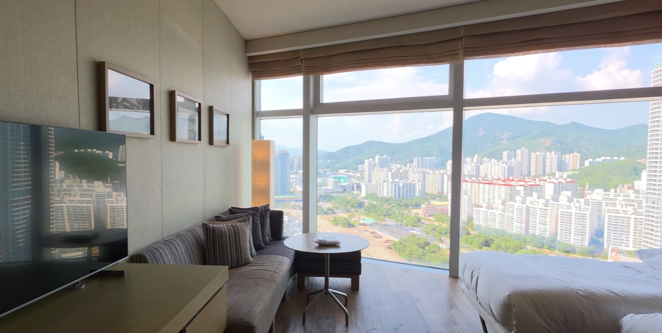 A sofa and table beside floor-to-ceiling windows, with city towers and hills outside a Park Hyatt Busan room.