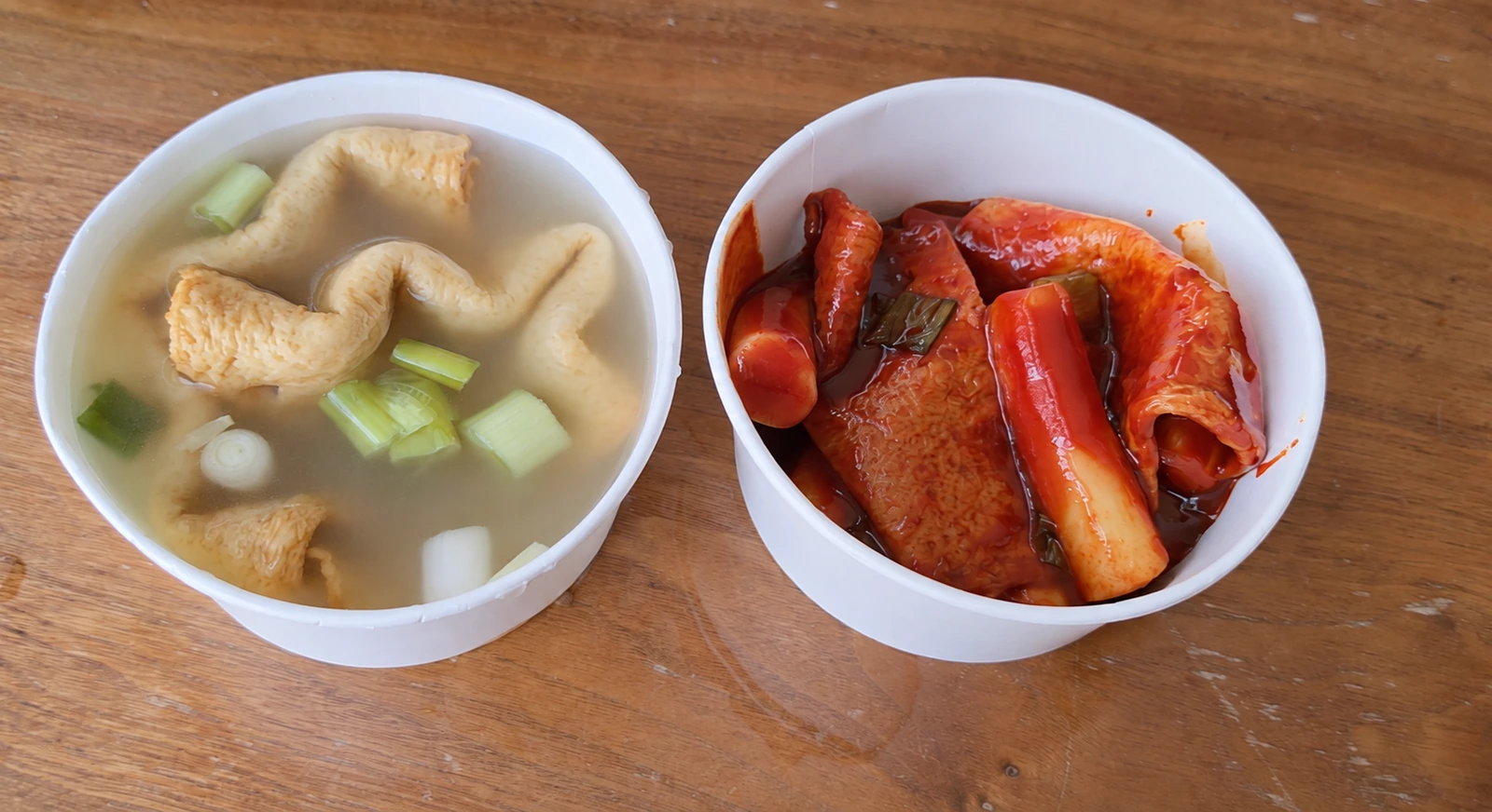Two white takeaway bowls containing red-sauce tteokbokki and fish-cake soup with sliced scallions.