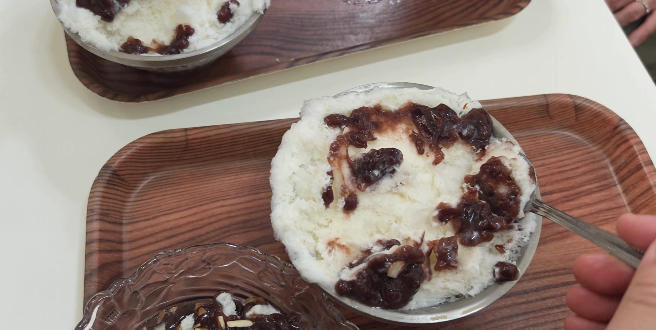 A bowl of white shaved ice topped with sweet red beans, served on a wood-grain tray.